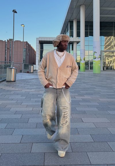 Cárdigan de punto beige con detalle "HOM", camiseta blanca, jeans claros desgastados y zapatillas beige; el modelo lleva un gorro de piel beige.