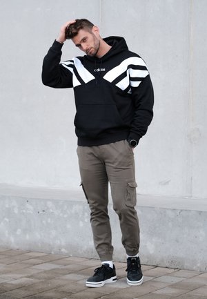 Sudadera negra con rayas diagonales blancas y logo, combinada con pantalones cargo oliva y zapatillas negras. El modelo tiene el cabello texturizado y lleva un reloj de pulsera.