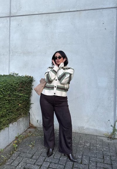 Mujer con cárdigan a rayas, pantalones negros y gafas de sol, posando con las manos cerca del rostro junto a un seto verde y una pared de hormigón gris.