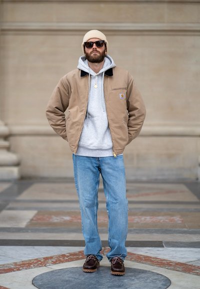 Chaqueta bomber marrón sobre una sudadera gris, jeans azul claro y zapatos marrones con cordones. Los complementos incluyen gafas de sol y un gorro beige.