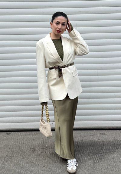 Blazer blanco ajustado sobre una blusa verde acanalada y una falda de satén oliva. Completo con un bolso acolchado de cadena y zapatillas blancas.