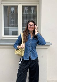 Denim button-up shirt with long sleeves, fitted style. Black track trousers with white stripes. Small yellow handbag with a rounded shape.