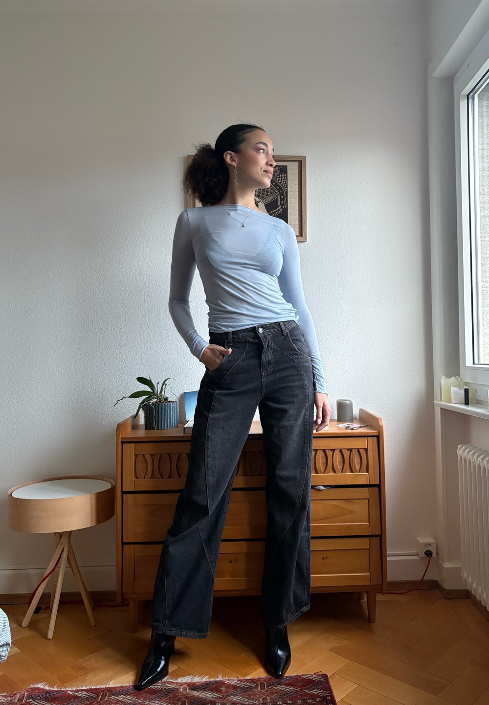 Femme aux cheveux bouclés attachés en queue de cheval, portant un haut à manches longues bleu clair et un jean noir à jambes larges, debout avec assurance à l'intérieur près d'une commode.
