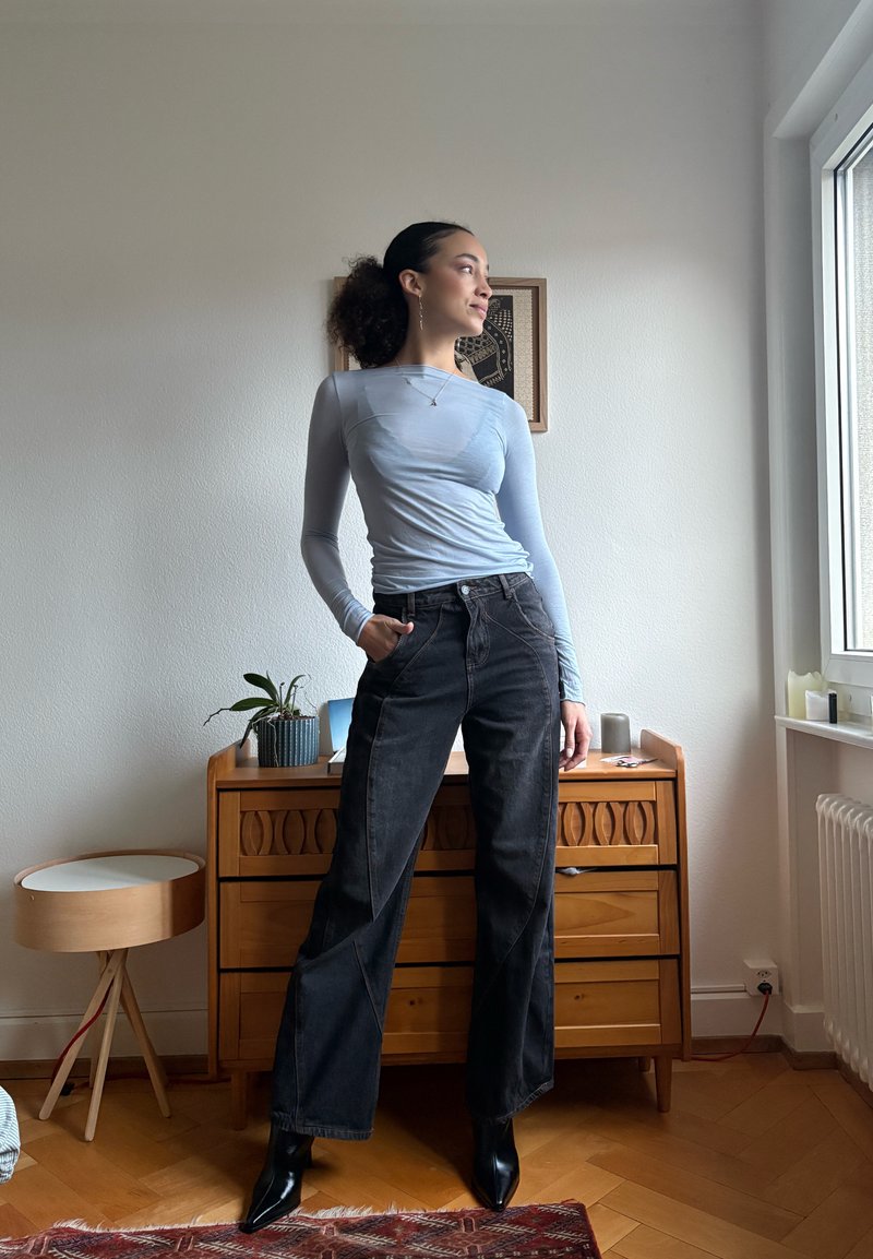 Femme aux cheveux bouclés attachés en queue de cheval, portant un haut bleu clair à manches longues et un jean large noir, debout avec assurance à l'intérieur près d'une commode.