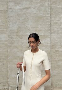 Abito color crema con maniche corte, tessuto texturizzato, bottoni frontali e silhouette aderente. Il modello tiene una tracolla bianca.