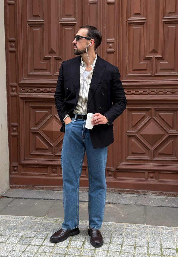 Hombre con gafas de sol y auriculares sostiene una taza de café, lleva una blazer negra, camisa de rayas, vaqueros azules y mocasines, de pie junto a una puerta marrón.