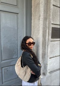 Bolsa de ombro em camurça bege, textura suave, forma hobo, grande abertura, combinada com uma jaqueta preta e óculos de sol oversized castanhos. Fundo de porta cinza.