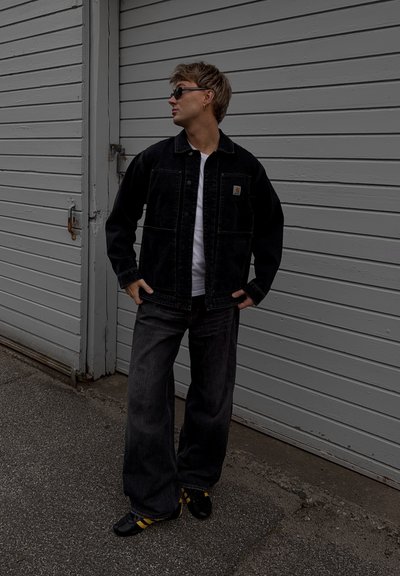 Chaqueta de denim negra sobre una camisa blanca, combinada con pantalones negros holgados y zapatos negros con rayas amarillas. De pie contra una pared gris.