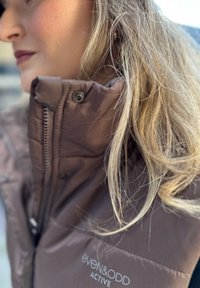 Hnědá prošívaná vesta s vysokým límcem, zipy a patentkami, s logem "even&odd ACTIVE". Lehký materiál s hladkou texturou.