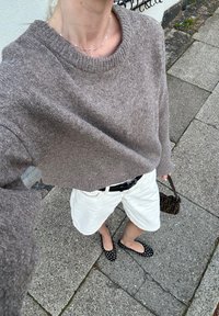 Πλεκτό καφέ πουλόβερ με στρογγυλή λαιμόκοψη, λευκές υφασμάτινες σορτς, μαύρα παπούτσια με πουά και μια μικρή τσάντα με σχέδιο. Ορατό τσιμεντένιο δάπεδο.