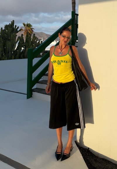 Top de tirantes amarillo con el texto "BRASIL", pantalones cortos atléticos negros con rayas blancas, zapatos de tacón negros y bolso de hombro oscuro. Escalera verde de fondo.
