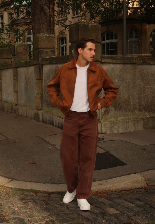 Giacca in suede marrone, t-shirt bianca, pantaloni larghi marroni e scarpe da ginnastica bianche. In piedi contro un muro di pietra, con pavimentazione e vegetazione nelle vicinanze.