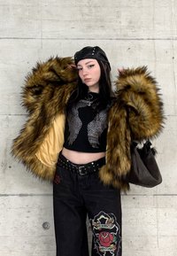 Νεαρός/ή ποζάρει μπροστά σε έναν τοίχο από σκυρόδεμα φορώντας καφέ γούνινο παλτό, μαύρο δερμάτινο καπέλο με καρφιά, μαύρο κοντό τοπ και μαύρο κεντητό παντελόνι.