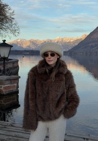 Femme en manteau de fourrure marron et chapeau beige debout près d'un lac avec des montagnes enneigées et un lampadaire en pierre au coucher du soleil.