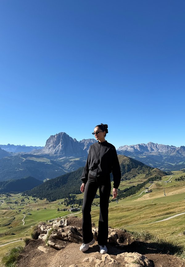 Woman wearing black clothes and sunglasses standing on a rocky hill overlooking a green valley and distant mountains under a clear blue sky.