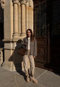 Károvaná bunda, krémový sveter, béžové džínsy a topánky na platforme v taupe farbe. Hnedá kabelka a pohár kávy. V pozadí sú kamenné stĺpy a drevené dvere.