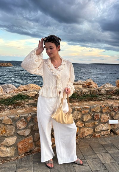 Blusa de un beige claro con cuello volantes y lunares; pantalones de tiro ancho blancos y texturizados; bolso pequeño dorado; sandalias grises; fondo costero.