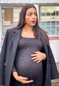 Abito in pizzo nero con scollo quadrato indossato sotto un cappotto nero. Capelli lunghi, smalto rosso e gioielli dorati valorizzano l'aspetto.