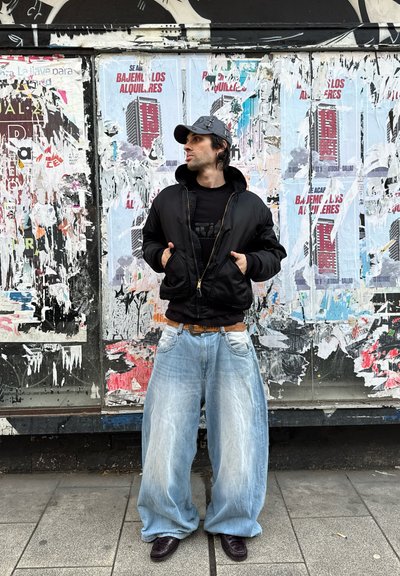 Chaqueta bomber negra, camiseta negra, jeans anchos de color azul claro, cinturón marrón y zapatos oscuros, contra un fondo de cartel colorido y desgastado.