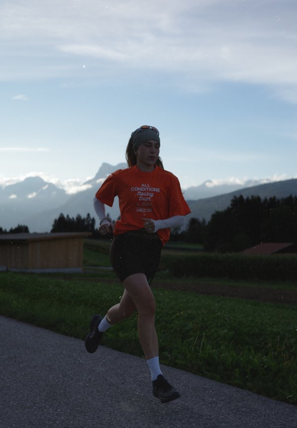 Un coureur en T-shirt orange trottine sur un chemin pavé, avec des champs verdoyants, une forêt et des montagnes en arrière-plan.