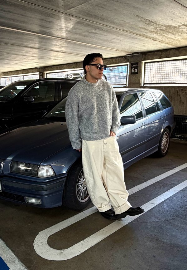 Uomo con maglione grigio e pantaloni bianchi si appoggia a una station wagon blu scuro in un parcheggio coperto al livello 6a.