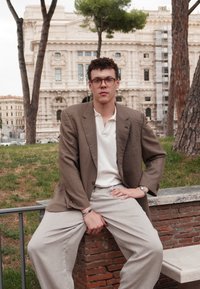 Blazer xadrez castanho sobre uma polo branca, combinado com calças de perna larga beija-flor claras. O fundo apresenta um edifício histórico e vegetação.