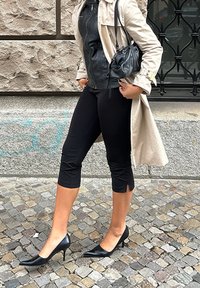 Cappotto beige, giacca di pelle nera aderente, leggings neri alla caviglia e tacchi a punta neri con una piccola borsa a tracolla. Sfondo in pietra testurizzata.