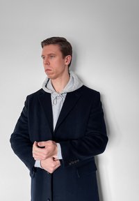 Άνδρας που φοράει γκρι φούτερ με κουκούλα κάτω από μαύρο παλτό, ρυθμίζοντας το μανίκι ενώ κοιτάζει προς τα αριστερά, μπροστά σε απλό ανοιχτογκρι φόντο.