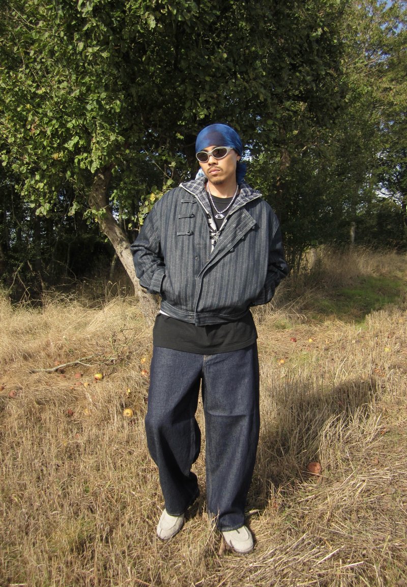 Un hombre con gafas de sol, un pañuelo azul en la cabeza, una chaqueta a rayas y vaqueros sueltos está de pie en un campo seco bajo los árboles.
