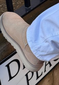 Botte en daim beige à semelle épaisse portée avec un pantalon bleu clair reposant sur une rambarde en métal noir au-dessus d'un panneau de rue indiquant "Dr Pla".