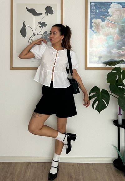 Blusa blanca corta con mangas abullonadas y detalle de cuerda, combinada con pantalones cortos negros. Accesorios incluidos: un bolso negro y tacones negros, calcetas blancas.