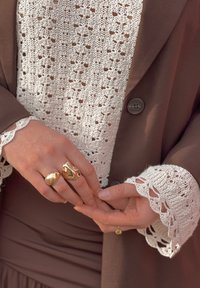 Blazer marron avec un chemisier blanc en maille orné d'un ourlet en dentelle. Mains décorées de deux bagues en or brillantes, l'une lisse et l'autre texturée.