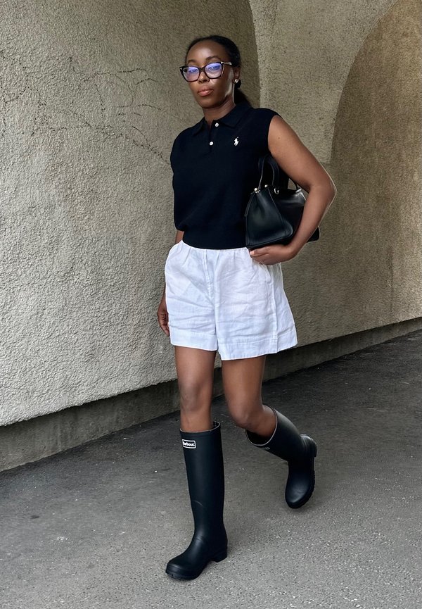 Femme portant un polo noir sans manches, un short blanc, des bottes de pluie noires et des lunettes, tenant un sac noir, marchant le long d'un mur texturé.