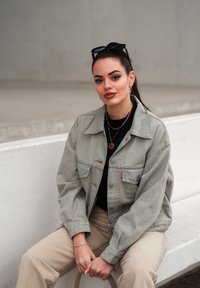 Světlá šedá džínová bunda s mírně oversized střihem, s knoflíkovým zapínáním a dvěma náprsními kapsami. Černý top a béžové kalhoty dokončují outfit.