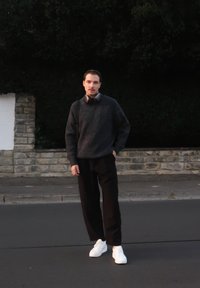 Sort sweater, mørke bukser og hvide sneakers. Iført over-ear høretelefoner, står på en gade med en stenvæg i baggrunden.