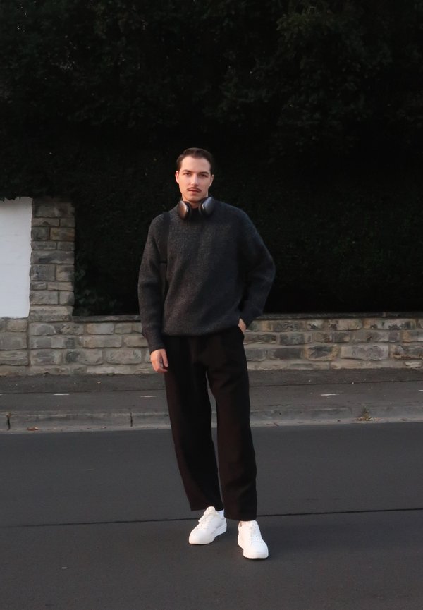 Schwarzer Pullover, dunkle Hose und weiße Sneaker. Trägt Over-Ear-Kopfhörer und steht auf einer Straße vor einer Steinmauer.