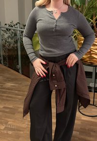 Cămașă henley gri cu mâneci lungi și cu o plachetă cu nasturi, purtată cu pantaloni negri cu picioare largi și o cămașă maro în carouri legată în talie.