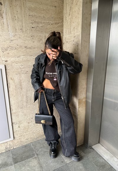 Mujer de pie junto al ascensor, con chaqueta de cuero negra, suéter marrón corto, pantalones anchos negros, botas negras y sosteniendo un bolso negro con cadena dorada.