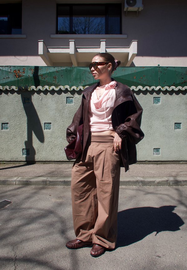 Žena oblečená v nadrozmernej hnedej koženej bunde, béžových širokých nohaviciach, ružovom tričku s kvetinovým detailom, slnečných okuliaroch, stojaca na slnkom zaliatej ulici.