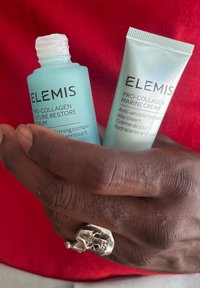 Due prodotti per la cura della pelle ELEMIS tenuti in una mano: una bottiglia di siero blu chiaro e un tubo di crema verde, entrambi con testo etichettato in nero.