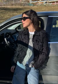 Texturierte schwarze Jacke mit einem flauschigen Design, kombiniert mit einer grauen Strickjacke, weißem Hemd und hellblauen Jeans. Stehend neben einem Auto.