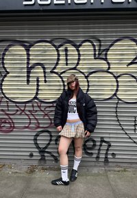 Casaco puffer preto, camisa curta branca com texto, saia de xadrez, meias brancas até ao joelho com riscas pretas, e sapatos de futebol pretos. Fundo urbano.