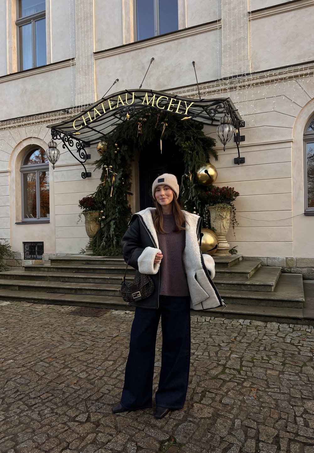 Casaco de shearling preto e creme, suéter cinza oversized, jeans escuros de perna larga e uma bolsa estampada, com degraus e vegetação festiva ao fundo.