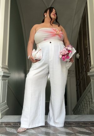 Pantalones anchos blancos de cintura alta con un botón frontal, combinados con un top sin tirantes que presenta un diseño floral rosa y sostiene un ramo.