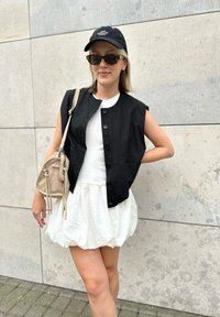 Svart ermeløs vest over en hvit topp, parret med et hvitt bobleskjørt. Tilbehøret inkluderer en svart caps og en beige veske med frynser.