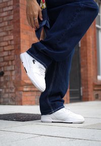 Bela teniska z gladkim usnjenim zgornjim delom, z luknjicami, ravnimi vezalkami in teksturiranim belim podplatom. V kombinaciji z ohlapnimi temno modrimi kavbojkami.