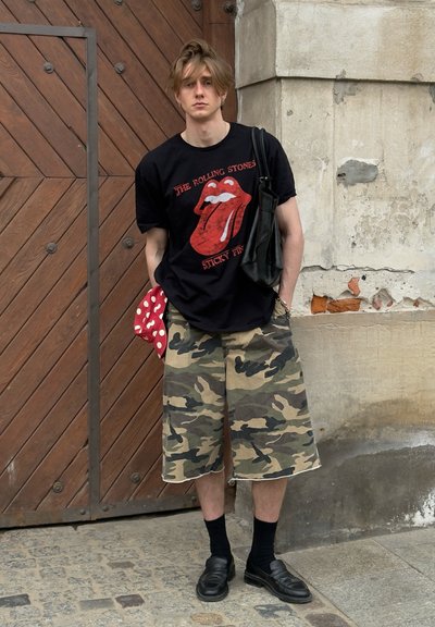 Joven con cabello despeinado que lleva una camiseta negra de los Rolling Stones, pantalones cortos de camuflaje, calcetines negros, mocasines y lleva dos bolsas, está junto a una puerta de madera.