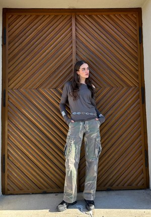 Top de manga larga gris con un cinturón de cadena plateada, combinado con pantalones cargo verde camuflaje y zapatillas negras, de pie frente a una puerta de panel diagonal de madera.