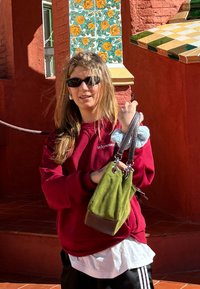Green suede handbag with brown leather accents and attached chain straps. The model is wearing a burgundy sweatshirt and black shorts, set against a background of colourful tiles.
