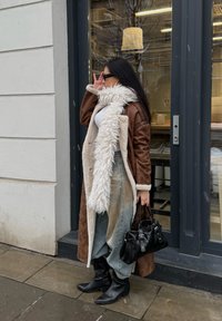 Rudos ir kreminės spalvos dirbtinio kailio paltas su pūstu apykakle, balta palaidinė, plačios šviesiai mėlynos džinsinės kelnės, juodi auliniai batai ir juoda rankinė.